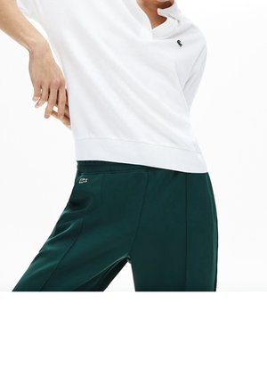 Polo shirt blanc avec col et un petit logo vert, associé à un pantalon lisse, vert foncé, fuselé avec des poches latérales.