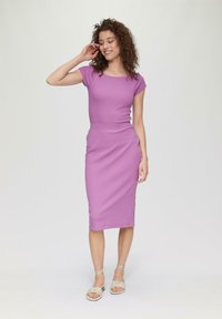 Paarse getailleerde jurk met een ronde hals en korte mouwen, tot op de knie, gecombineerd met lichtgekleurde sandalen met bandjes, op een model met krullend haar.