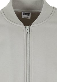 Giacca zip-up grigia chiara con colletto a coste e tessuto testurizzato. Presenta una zip frontale con denti in argento. Etichetta visibile all'interno.