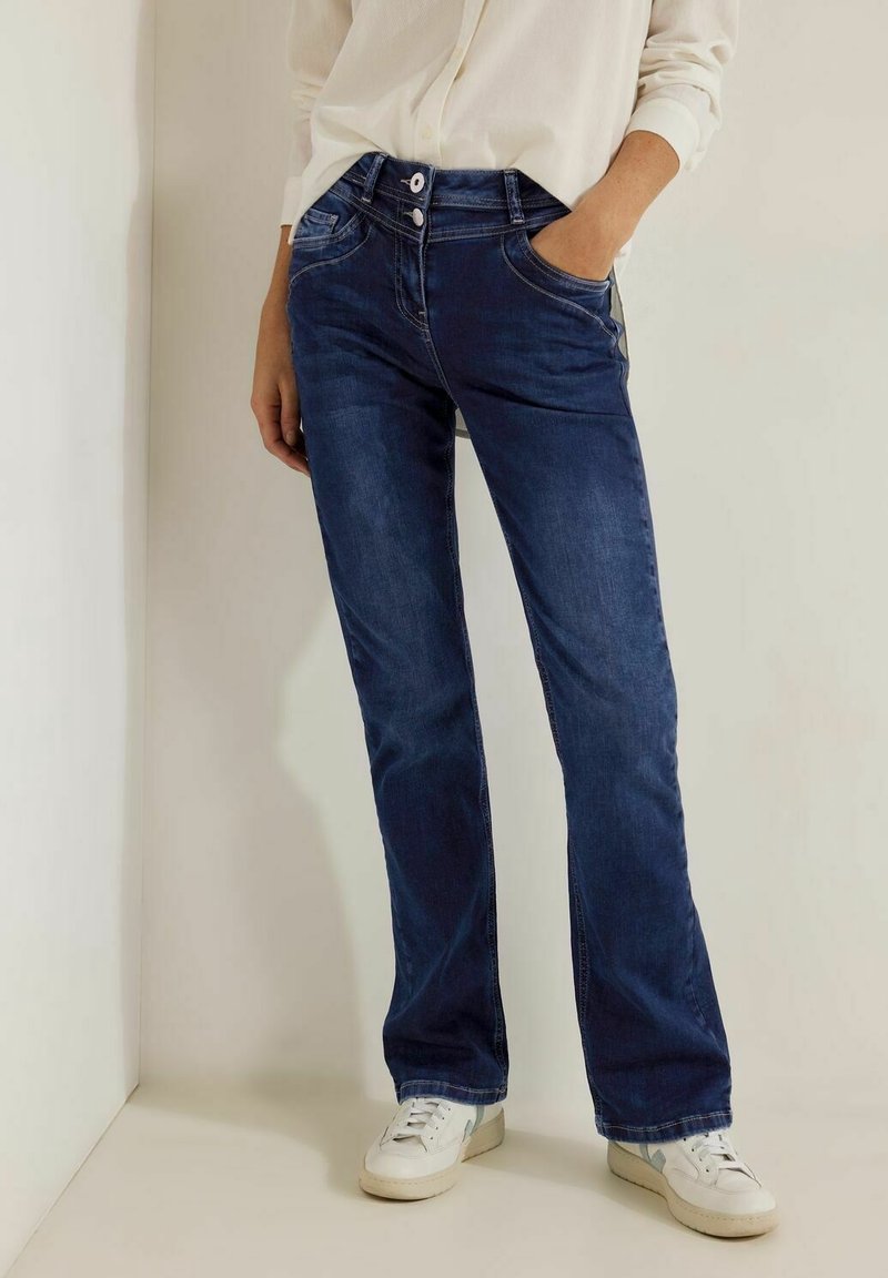Jeans de mezclilla azul oscuro con forma acampanada, que cuentan con bolsillos frontales y un cierre de doble botón. Textura suave y detalles mínimos.