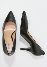 Escarpins noirs en cuir verni à talons hauts avec un design à bout pointu. Les chaussures présentent une finition lisse et un logo discret sur la semelle intérieure.