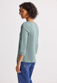 Vrouw met kort, krullend haar, gekleed in een lichtgroen shirt met lange mouwen en blauwe jeans, die zijwaarts staat tegen een effen achtergrond.
