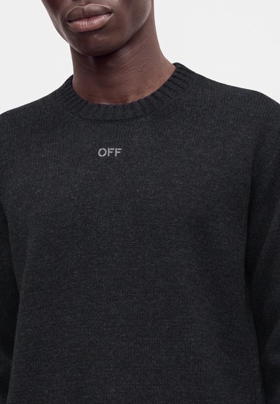 Jeune personne portant un pull en maille foncé avec col rond côtelé et le mot "OFF" brodé en lettres claires sur la poitrine.