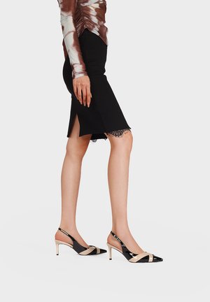 Donna con gonna a matita nera al ginocchio con orlo in pizzo e spacco, top a maniche lunghe con motivo, indossa décolleté slingback nere e beige con tacco basso.