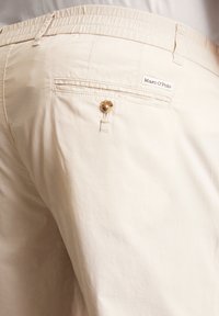Beige katoenen broek met een elastische tailleband, voorzien van een enkele achterzak met een knoop en een klein merkembleem.