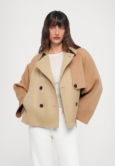 Double-breasted tan wool blend jacket with large buttons, extending sleeves, and a structured collar. White top worn underneath.
