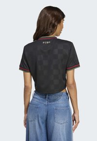 Donna con i capelli castani di lunghezza media che indossa una maglietta nera corta con bordo rosso e "FCBM" sulla schiena, abbinata a jeans blu a vita alta.