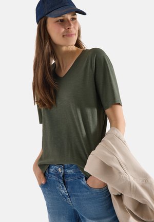 Jonge vrouw die een donkergroene V-hals T-shirt en blauwe spijkerbroek draagt, een beige jas vasthoudt en een donkerblauwe baseballpet draagt.