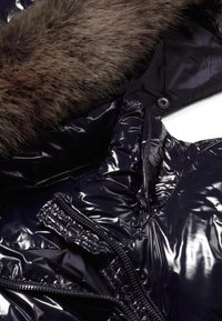 Chaqueta tipo plumón en un oscuro color marino con acabado brillante; presenta un cuello forrado de piel, detalles de cremallera y una textura suave y acolchada.