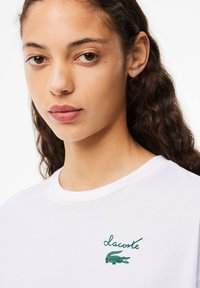 Baltos apvalios iškirptės marškinėliai su žalia išsiuvinėta "Lacoste" logotipo ir krokodilo simbolika. Lygi tekstūra, atsipalaidavęs siluetas.