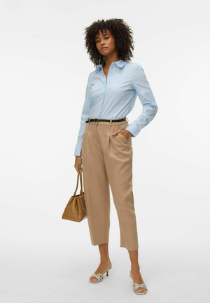 Femme debout portant une chemise bleu clair, un pantalon beige raccourci, portant un sac à main marron et des sandales blanches à talons sur un fond blanc.