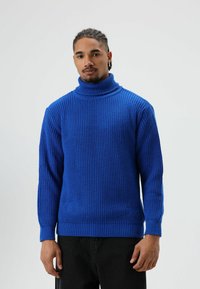 Blauwe geribbelde gebreide coltrui met lange mouwen, met een hoge kraag en een gestructureerde stof. Gecombineerd met donkere broek.