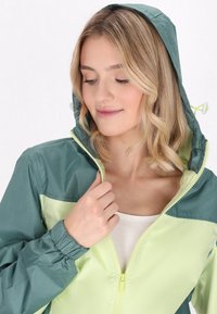 Chaqueta ligera con un diseño bicolor en verde y amarillo pálido. Incluye capucha, puños elásticos y un cierre frontal con un tirador en contraste.