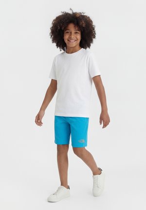 Enfant souriant aux cheveux bouclés portant un t-shirt blanc, un short bleu vif et des baskets blanches, debout devant un fond uni.