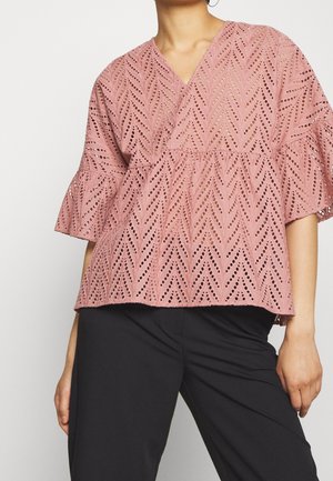 Femme portant un chemisier rose à motif, manches courtes évasées, et un pantalon noir sur un fond uni.