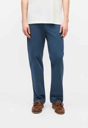 Mand iført blå bukser med lige ben, brune læder bådsko og en hvid t-shirt, stående mod en ensartet lys baggrund.