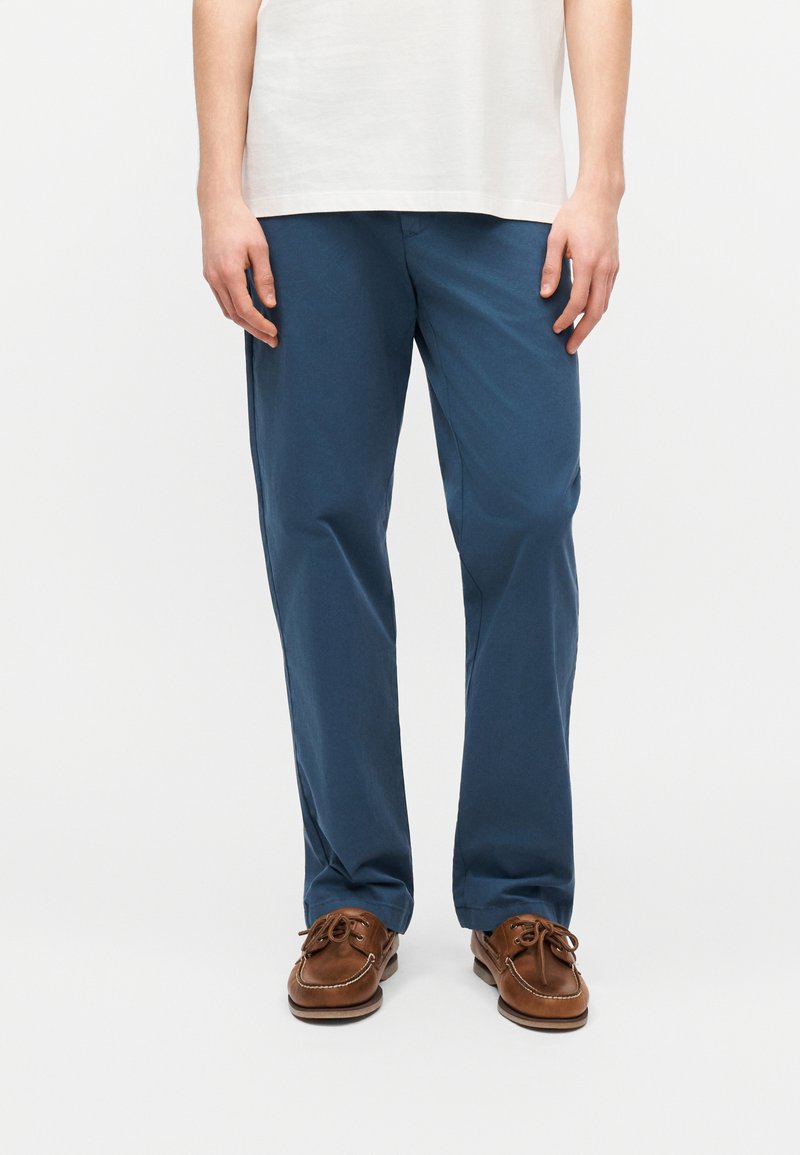 Άνδρας που φοράει μπλε παντελόνι με ίσια γραμμή, καφέ δερμάτινα παπούτσια σκαφάτου τύπου και λευκό μπλουζάκι, στέκεται μπροστά σε μια απλή ανοιχτόχρωμη επιφάνεια.