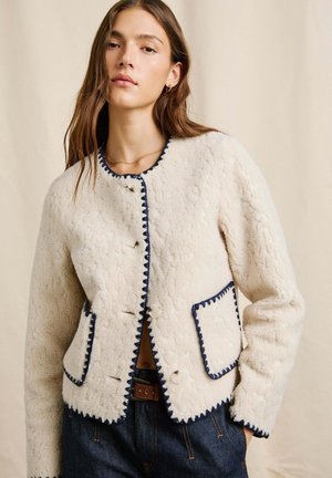 Mīksts krēmkrāsas žakets, izgatavots no šerlinga, ar tumši zilu apdari ap malām un pogām. Divas priekšējās kabatas ar pāršūtu detaļu.