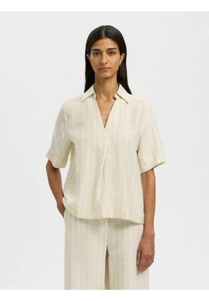 Žena s dlhými tmavými vlasmi má na sebe krémovú blúzku s krátkym rukávom a golierom a k nej ladúce široké nohavice, stojí pred jednoduchým svetlým pozadím.