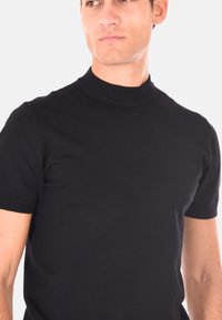 Uomo che indossa una maglietta a maniche corte nera aderente in maglia con colletto a coste, che guarda di lato su uno sfondo chiaro e semplice.
