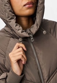 Ruda siūta striukė su gobtuvu, užsegama užtrauktuku ir su raišteliais. Charakterizuoja lygi tekstūra ir ribinėmis rankogaliais, pabrėžianti šiuolaikinį dizainą.