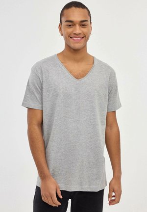Camiseta de algodón gris con escote en V, mangas cortas, dobladillo recto y una textura suave, llevada por una persona de pie contra un fondo neutro.