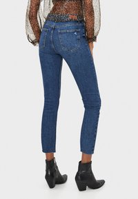 Jeans de mezclilla azul de talle alto, con un ajuste slim y longitud recortada, que presentan un sutil desvanecimiento y dos bolsillos traseros. Se combinan con botas negras hasta el tobillo.