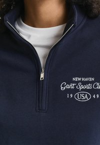 Close-up van een marineblauwe sweater met ritssluiting en de tekst "New Haven Gant Sports Club 1949 USA" geborduurd op de borst, gedragen over een wit shirt.