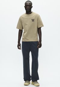 T-shirt in cotone beige con maniche corte e un piccolo logo nero sul petto, abbinato a pantaloni jeans scuri a vestibilità rilassata e scarpe beige.