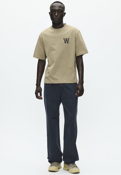 T-shirt en coton beige à manches courtes avec un petit logo noir sur la poitrine, associé à un pantalon décontracté bleu foncé et des chaussures beiges.