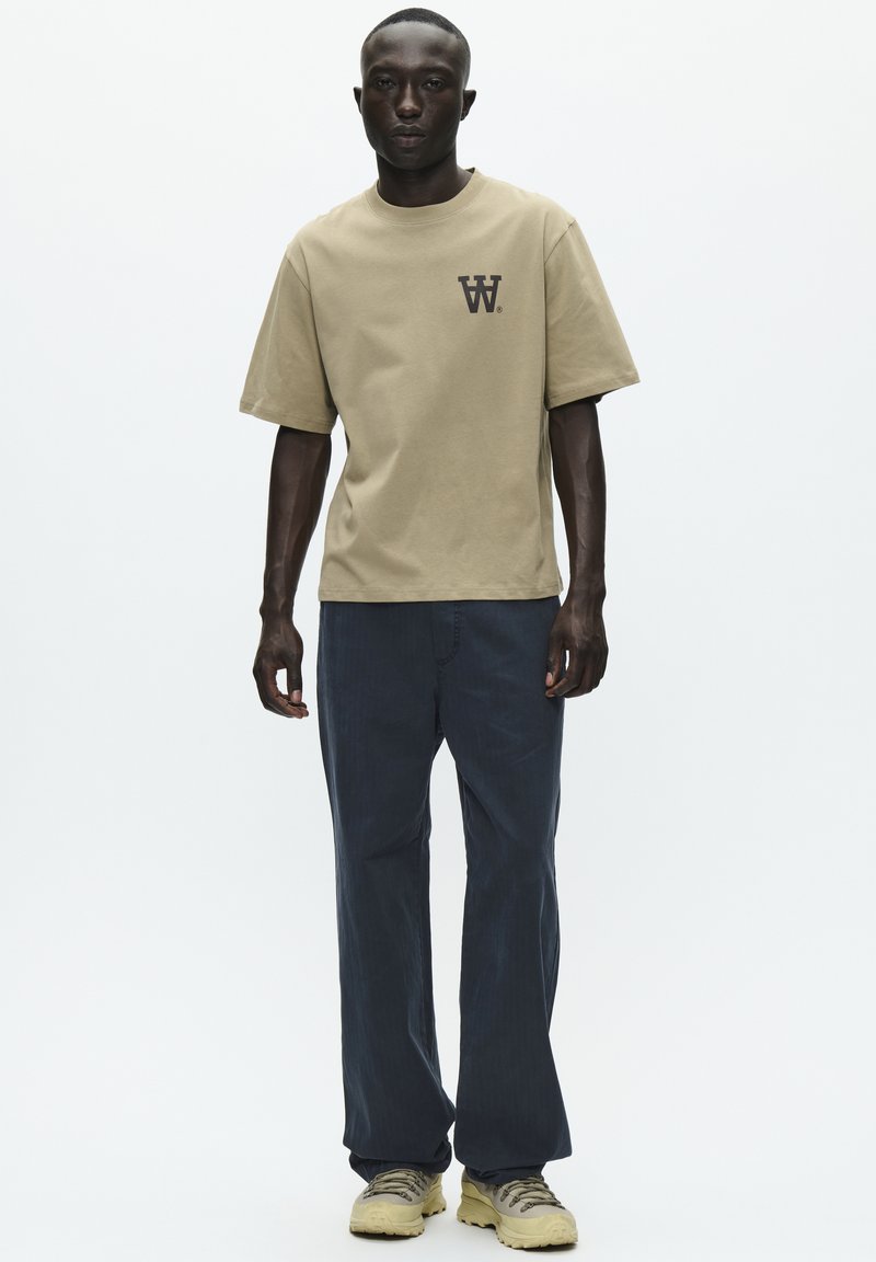 Camiseta de algodón beige con mangas cortas y un pequeño logo negro en el pecho, combinada con pantalones de corte relajado azul oscuro y zapatos beige.