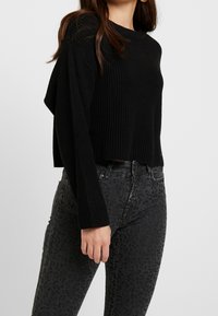 Svart ribbat kort tröja med långa ärmar, ihop med mörkgrå jeans med leopardmönster, som visar en texturerad tyg och figursydd design.