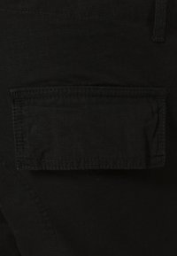 Black fabric with a textured surface features a rectangular patch pocket on the hip, outlined by contrasting stitching.