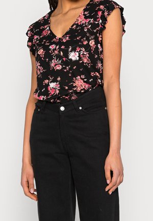 Kvinde iført en sort blomstret V-hals top med flæseærmer, der er stoppet ned i højtaljede sorte bukser mod en ensfarvet baggrund.