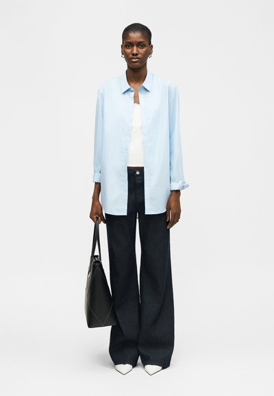 Femme grande portant une chemise oversize bleu clair sur un top blanc, pantalon large noir, chaussures blanches pointues, tenant un sac cabas noir, debout devant un fond uni.