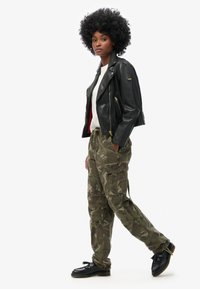 Zwarte leren motorjack met gouden hardware, witte t-shirt, camouflage cargo-broek en zwarte schoenen, model staand tegen een effen achtergrond.