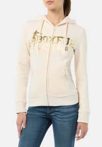 Beige Kapuzenjacke mit Reißverschluss, goldbedrucktem Text, Kordelzugkapuze, Vordertaschen sowie gerippten Bündchen und Saum. Weiche Stoffstruktur.