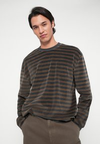 Νεαρός άνδρας με μεσαίου μήκους σκούρα μαλλιά, φορώντας καφέ και μαύρη ριγέ μπλούζα με μακριά μανίκια και καφέ παντελόνι, με τα χέρια στις τσέπες, σε ουδέτερο φόντο.