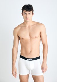 White cotton boxer shorts with a black elastic waistband displaying the brand "BOSS." Model poses with a neutral background.