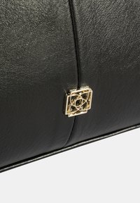 Zwarte leren tas met een gestructureerde afwerking, voorzien van een gouden geometrisch embleem als opvallend ontwerpelement aan de voorkant.