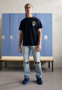 Črn bombažni t-shirt z grafičnim dizajnom, svetlo modri razorovani kavbojki in črne teniske z modrimi poudarki, stoji pred omarami.