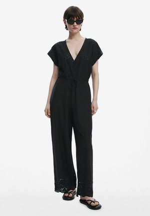 Black jumpsuit with a deep V-neck, short sleeves, and a drawstring waist. Features lace detailing at the hem and loose, wide legs.