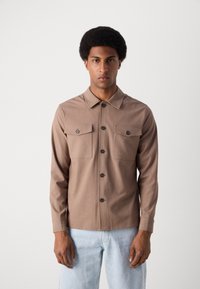Camisetă lungă, maro, cu mânecă lungă, dotată cu două buzunare pe piept, închidere cu nasturi și guler. Confecționată dintr-un material neted, cu o croială ajustată.