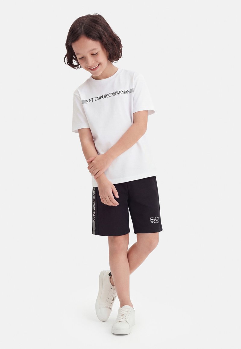 Enfant souriant aux cheveux courts et foncés portant un t-shirt blanc EA7 Emporio Armani, un short noir et des baskets blanches, debout avec les jambes croisées.