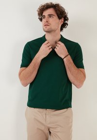 Zelený polo tričko vyrobené z hladkej látky, s golierom a krátkymi rukávmi. Model si upravuje výstrih, na sebe má béžové nohavice.