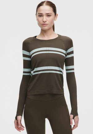 Kobieta o brązowych włosach spiętych w kucyk, ubrana w oliwkową bluzkę z długim rękawem z jasnoniebieskimi poziomymi paskami oraz dopasowane legginsy, stoi prosto.