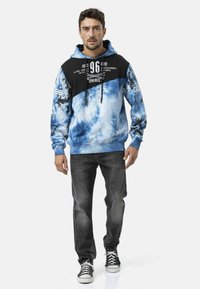 Blauwe tie-dye hoodie met een zwart bovenstuk, voorzien van grafische prints en trekkoorden. Gecombineerd met donkeren spijkerbroek en sneakers.