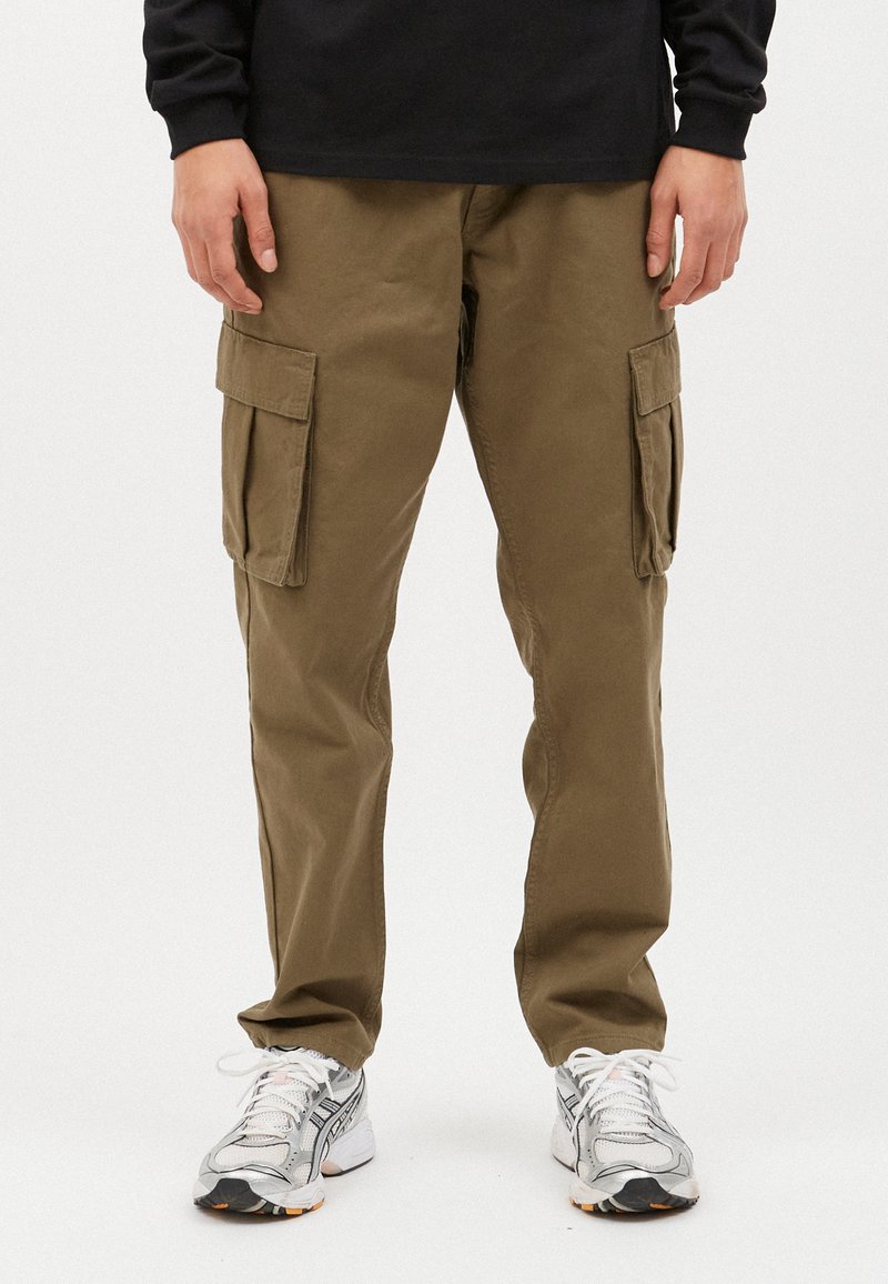Khaki Cargo-Hosen mit mehreren Taschen, aus Baumwollstoff. Getragen mit grauen und weißen Sneakers mit schwarzen und orangefarbenen Akzenten.