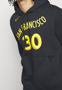 Sweat à capuche noir avec le texte jaune "SAN FRANCISCO 30" et le swoosh Nike jaune porté par une personne aux cheveux bouclés, vue en partie.