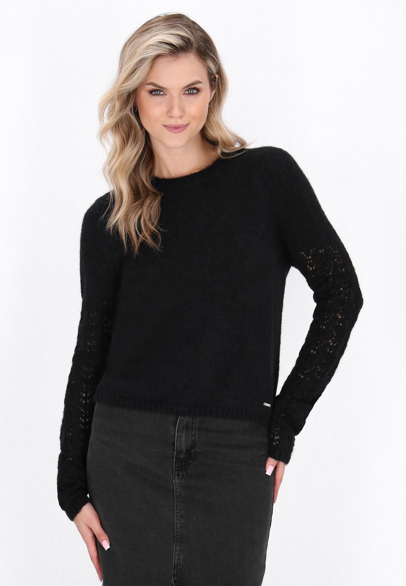 Femeie blondă îmbrăcată într-un pulover negru din tricot cu mâneci din dantelă și o fustă din denim închis, pozând cu o mână pe șold, pe un fundal alb.